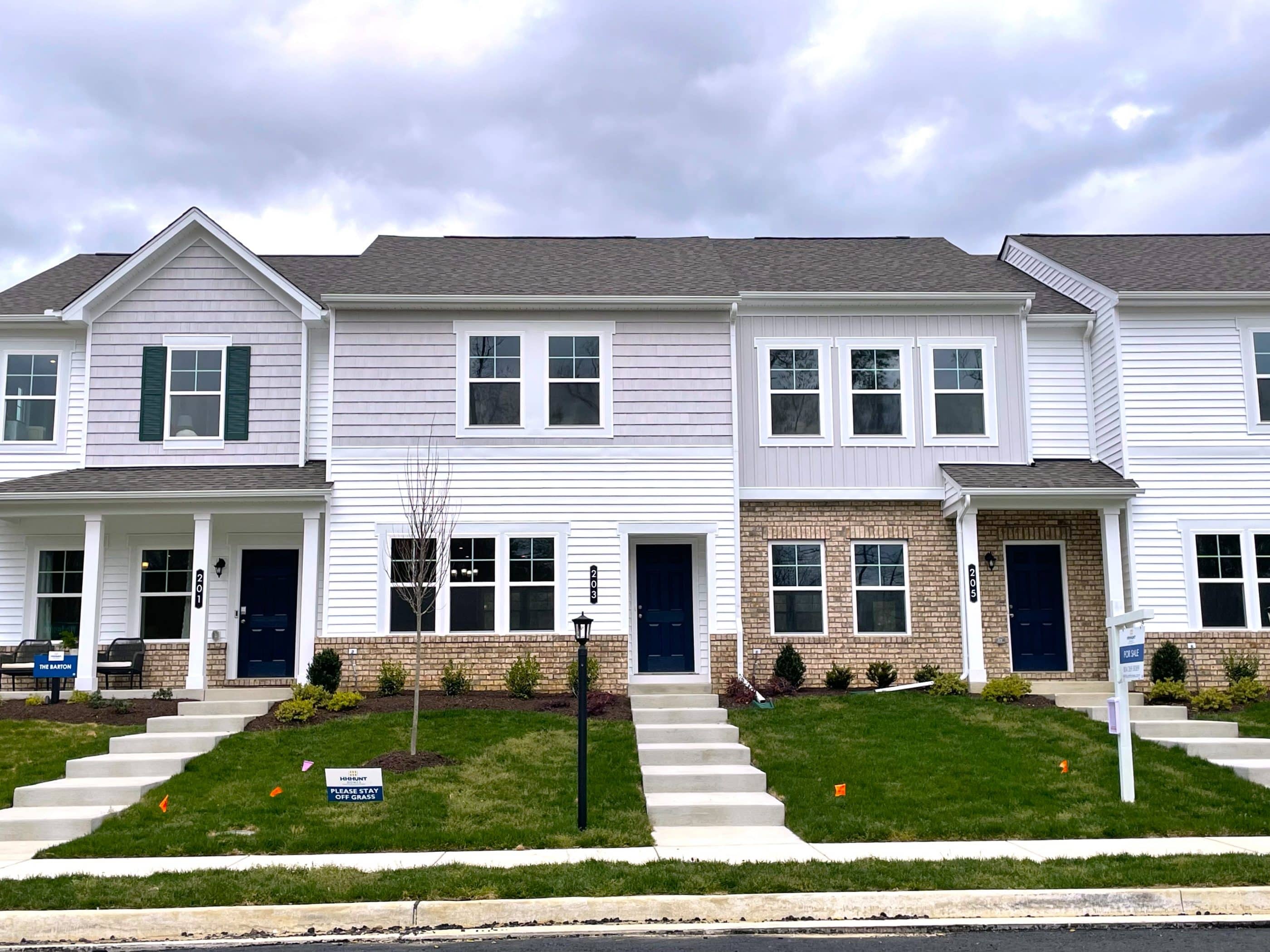 A front view of three connected residential homes in Virginia. The homes are white and light-gray, and some have decorative brick detailing. A set of stairs leads to the front door of each house, and each has a small front lawn. A sidewalk can be seen in the foreground.