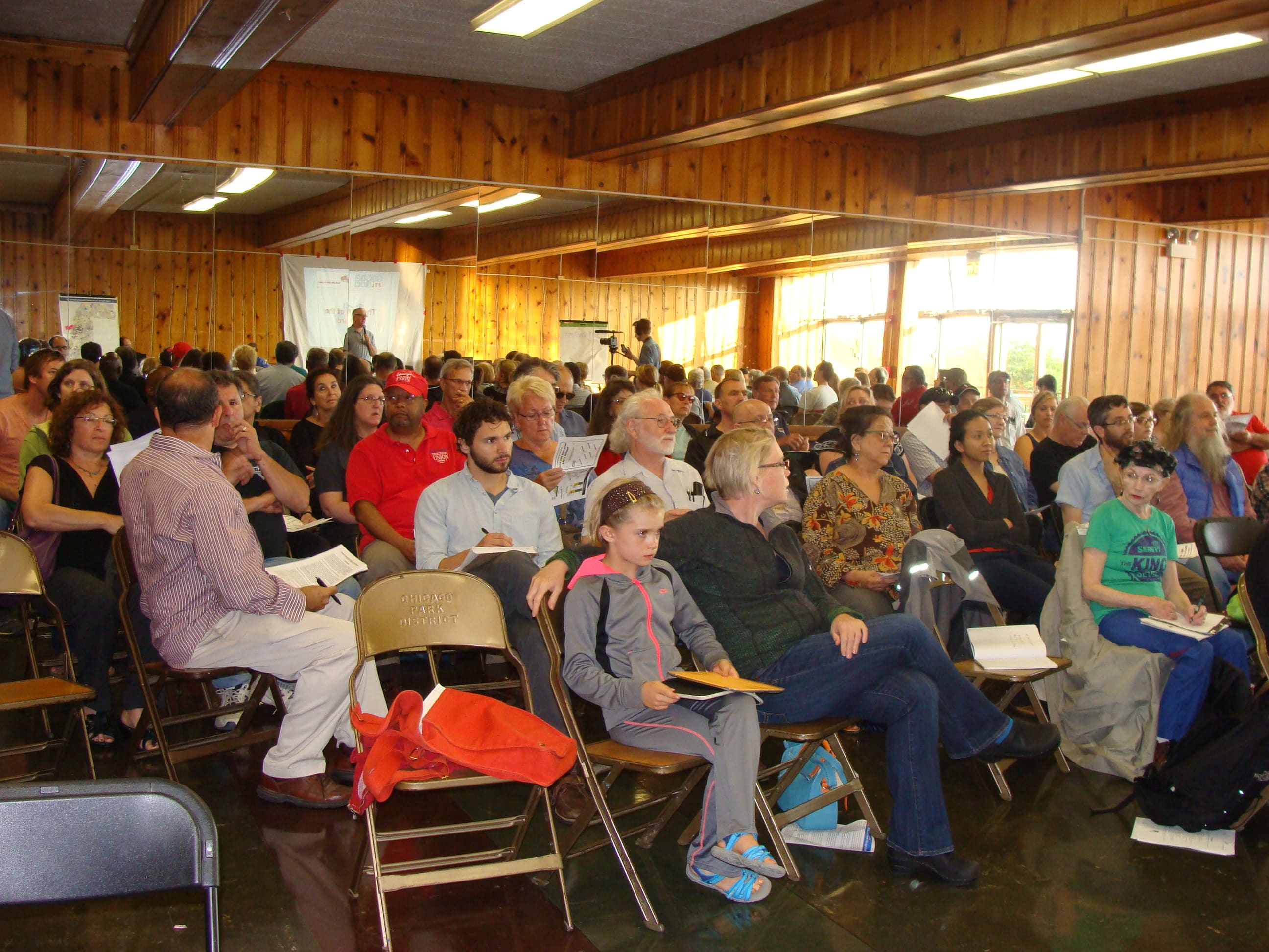 A large group of people of all different ages, races, and genders sit on folding chairs in what appears to be a community room. Many people can be seen holding pamphlets or notebooks. The room is paneled in wood and has a large full-wall mirror on the back wall.