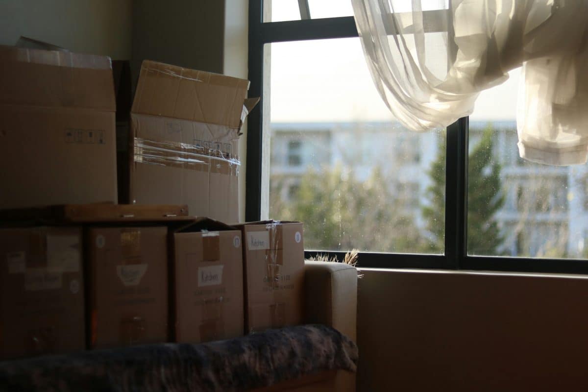 The interior of an apartment, showing a stack of moving boxes on top of a couch. Next to the couch is a large window with what appears to be an apartment building in the background.