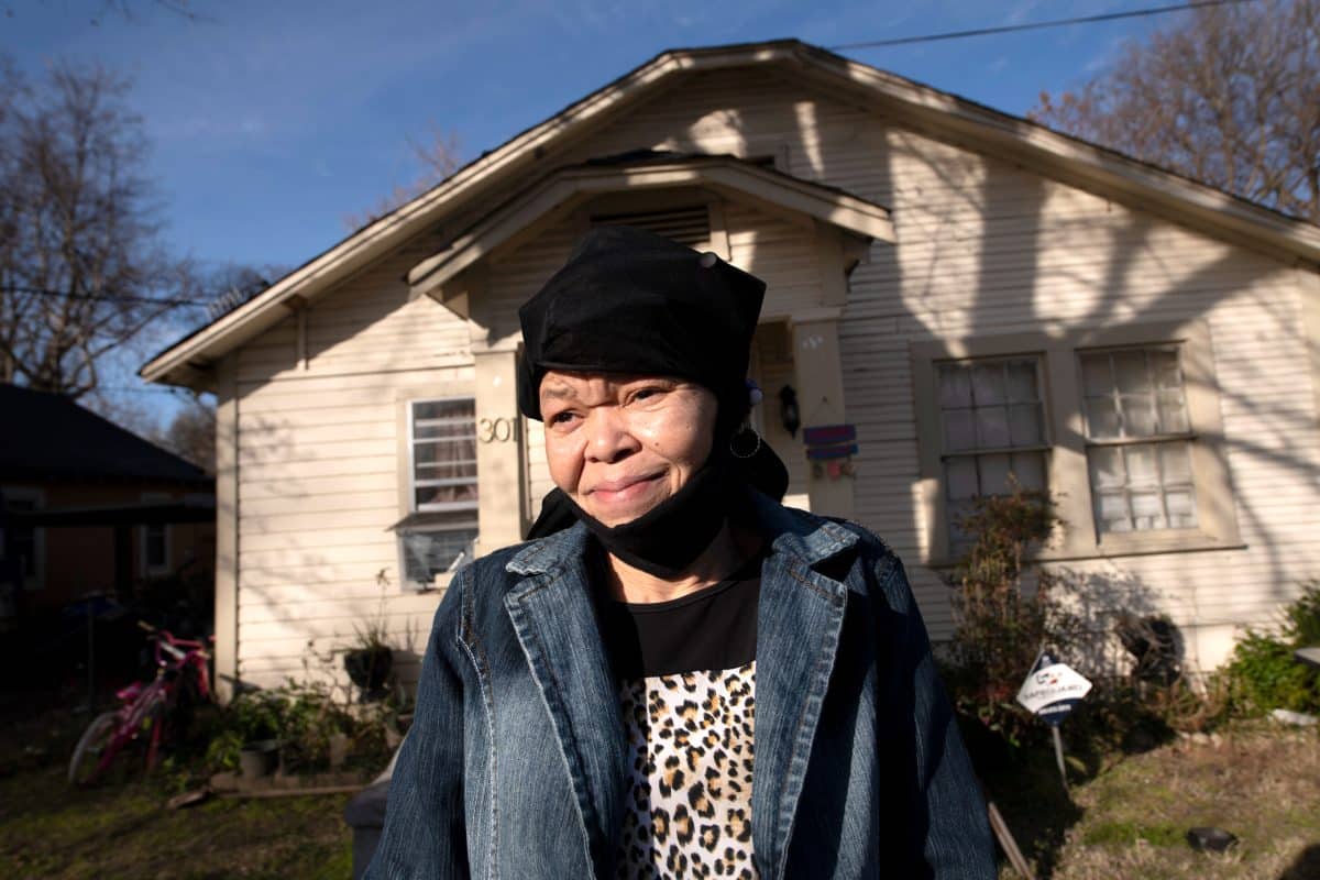 A middle-aged woman wearing a cheetah print shirt, a denim blazer, and a black hair covering or hat, standing in front of a small, beige-colored home.