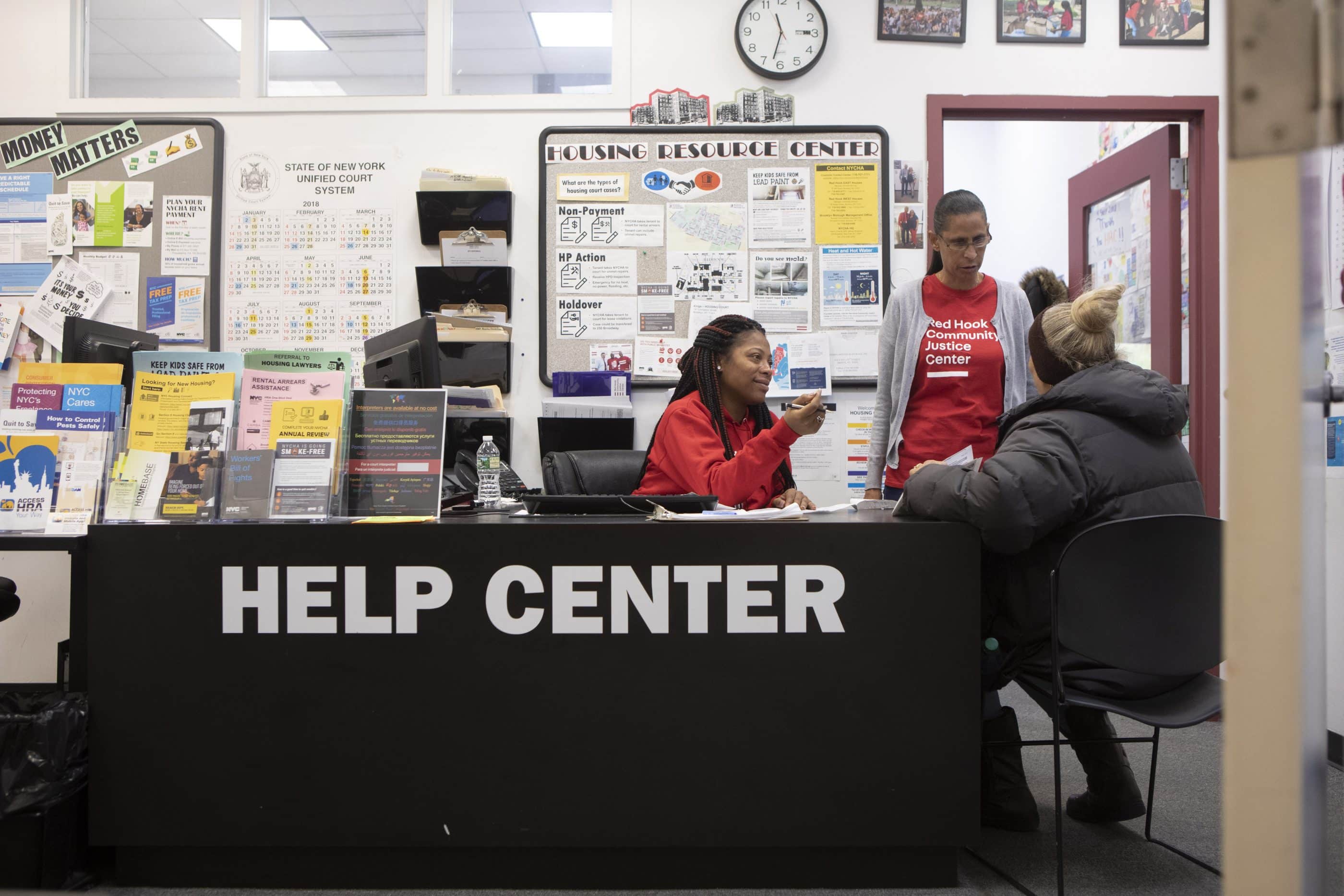 A black woman with braided hair and a red sweatshirt sits at a help center desk. Another woman stands next to her, wearing a red-and-white shirt that reads "Red Hook Community Justice Center." A woman with her back to the camera sits on a chair beside the desk; she is wearing a black puffer jacket and a knitted headband. Large signs saying "Housing Resource Center," "State of New York Unified Court System," and more are displayed behind the help center desk. Many different pamphlets on housing and workers' rights are displayed on the desk.