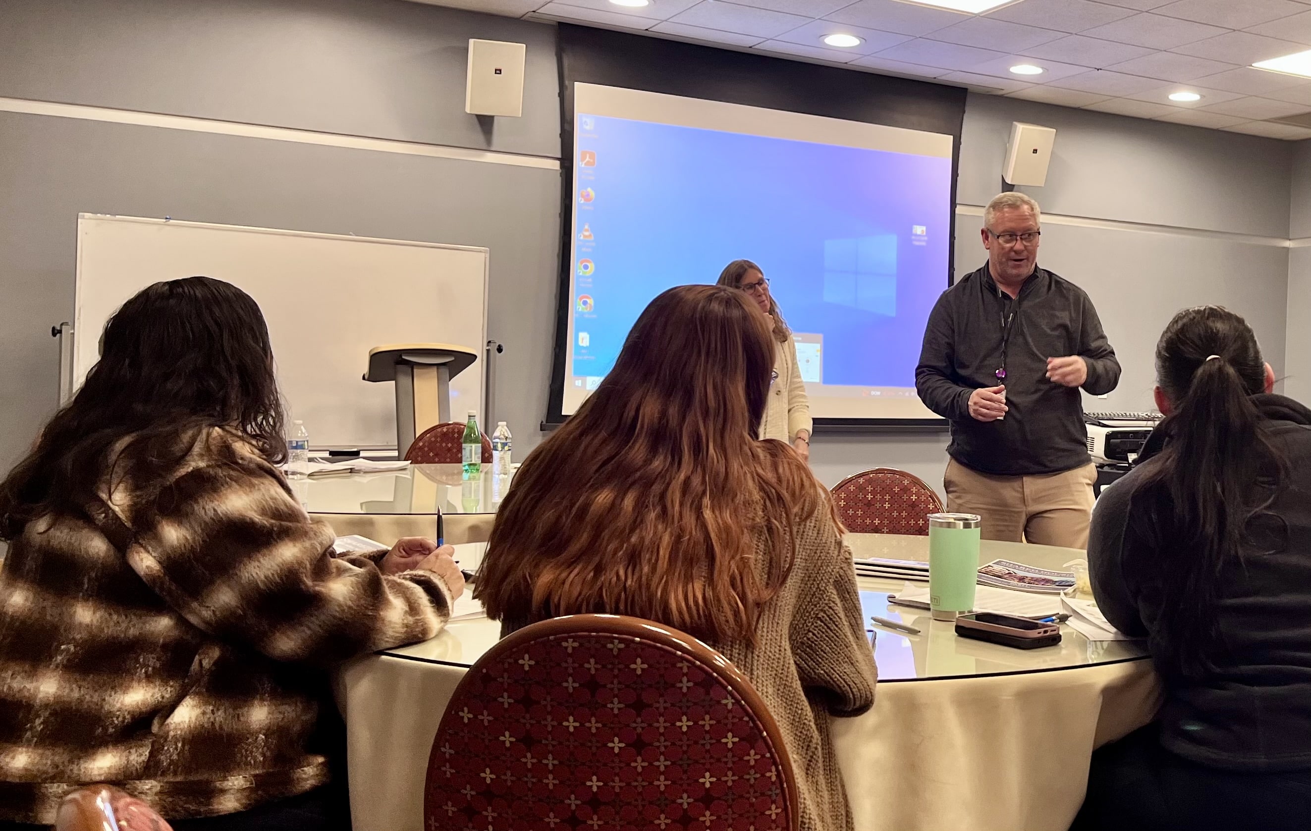 A group of people listen to a man who is standing in front of a projector screen.
