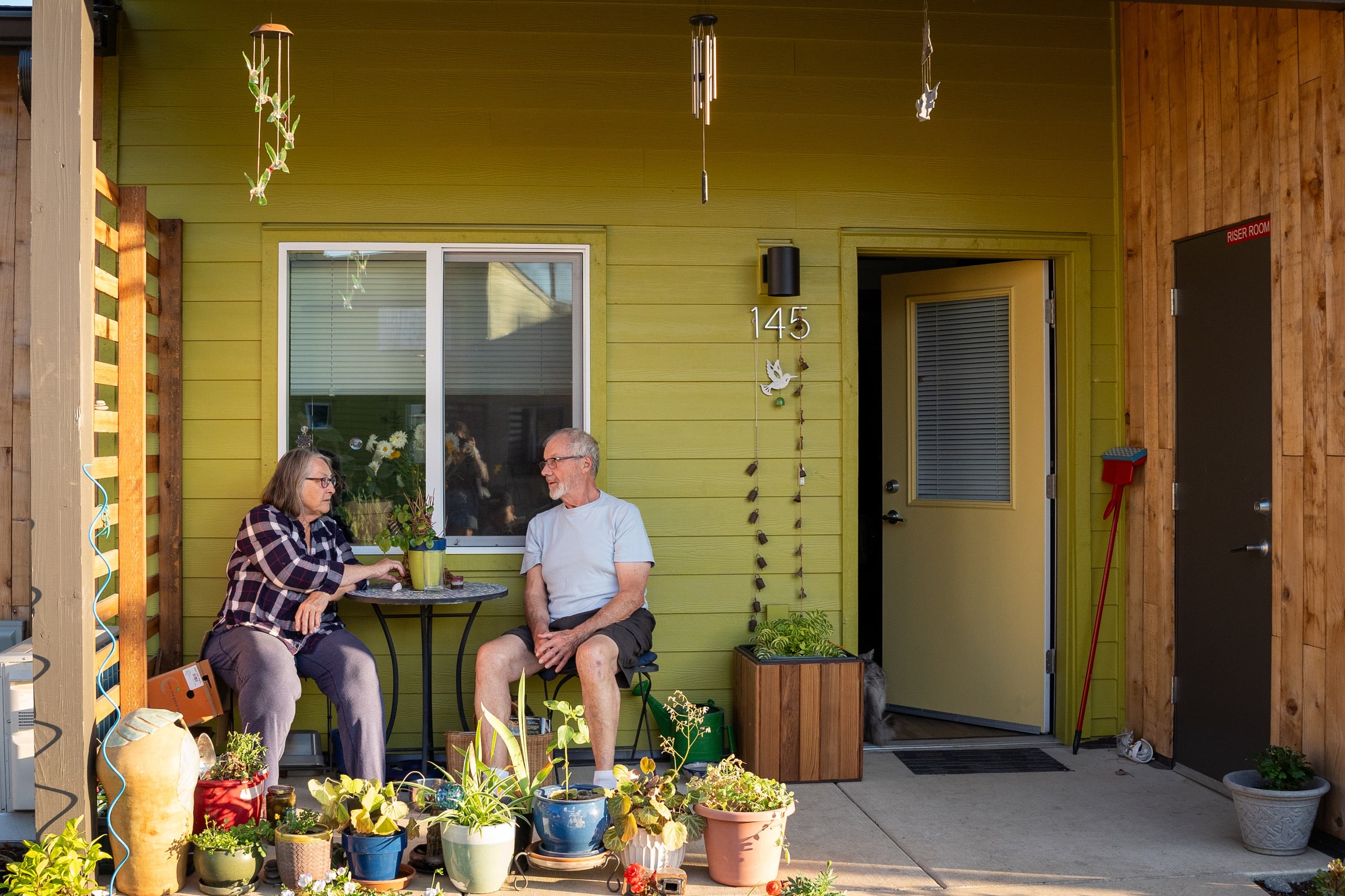 A woman wearing a black-and-white plaid shirt and gray pants sits at a small bistro table on a front patio with a man wearing a light-gray t-shirt and dark-gray cargo shorts. The patio is attached to the front of a green-colored house marked 145. On the patio are many potted plants, decorative wind chimes, and gardening tools. The front door of the house is slightly ajar, and a cat can be seen walking into the home.