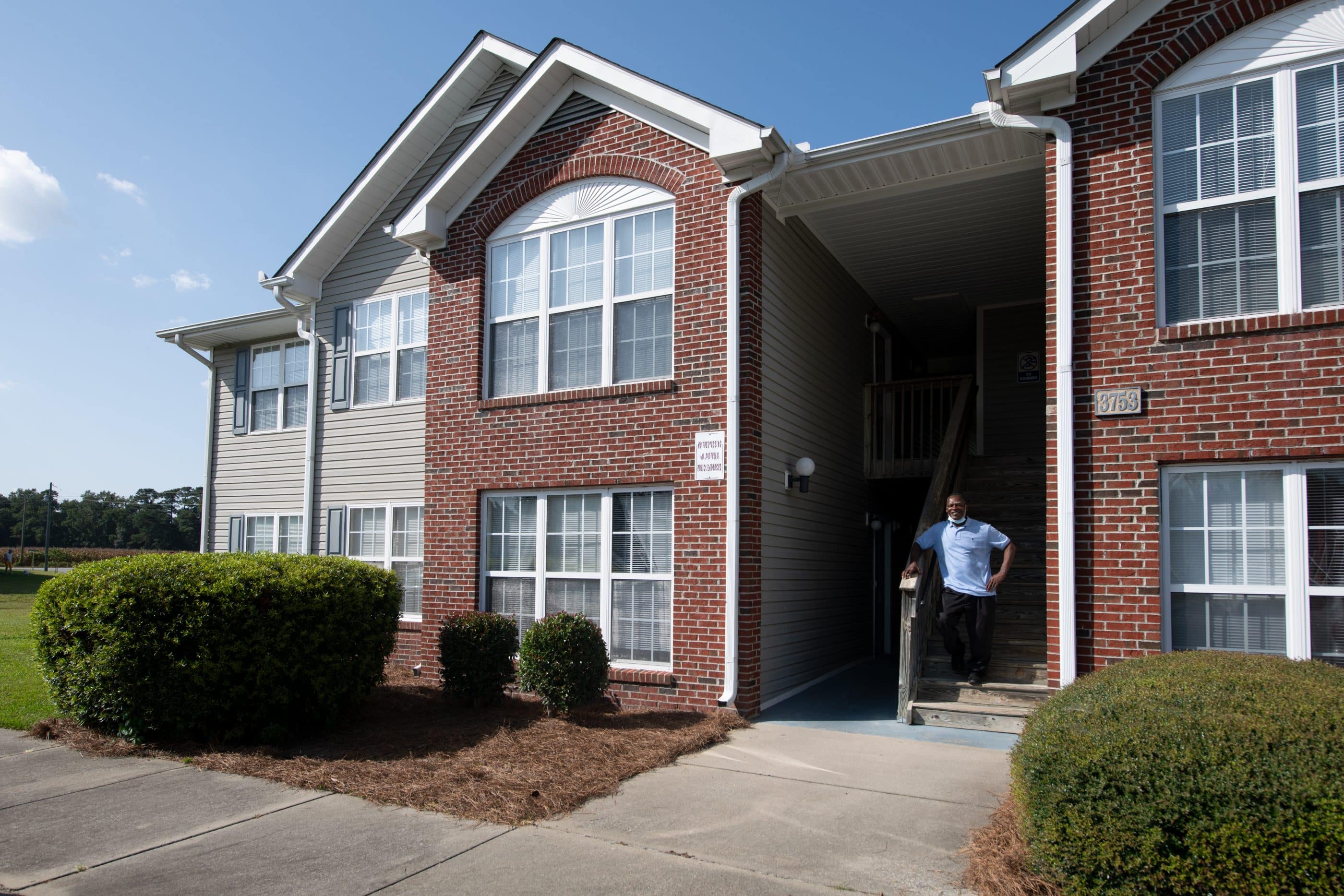 A middle-aged Black man wearing a blue polo shirt and black pants stands on a wooden staircase outside of a large brick multifamily home. In front of the home are several low shrubs and a pedestrian walkway.