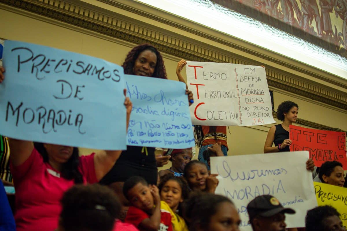 A group of people—mainly women and children—hold signs with Portuguese writing. Some of the signs say "Termo Territorial Coletivo."