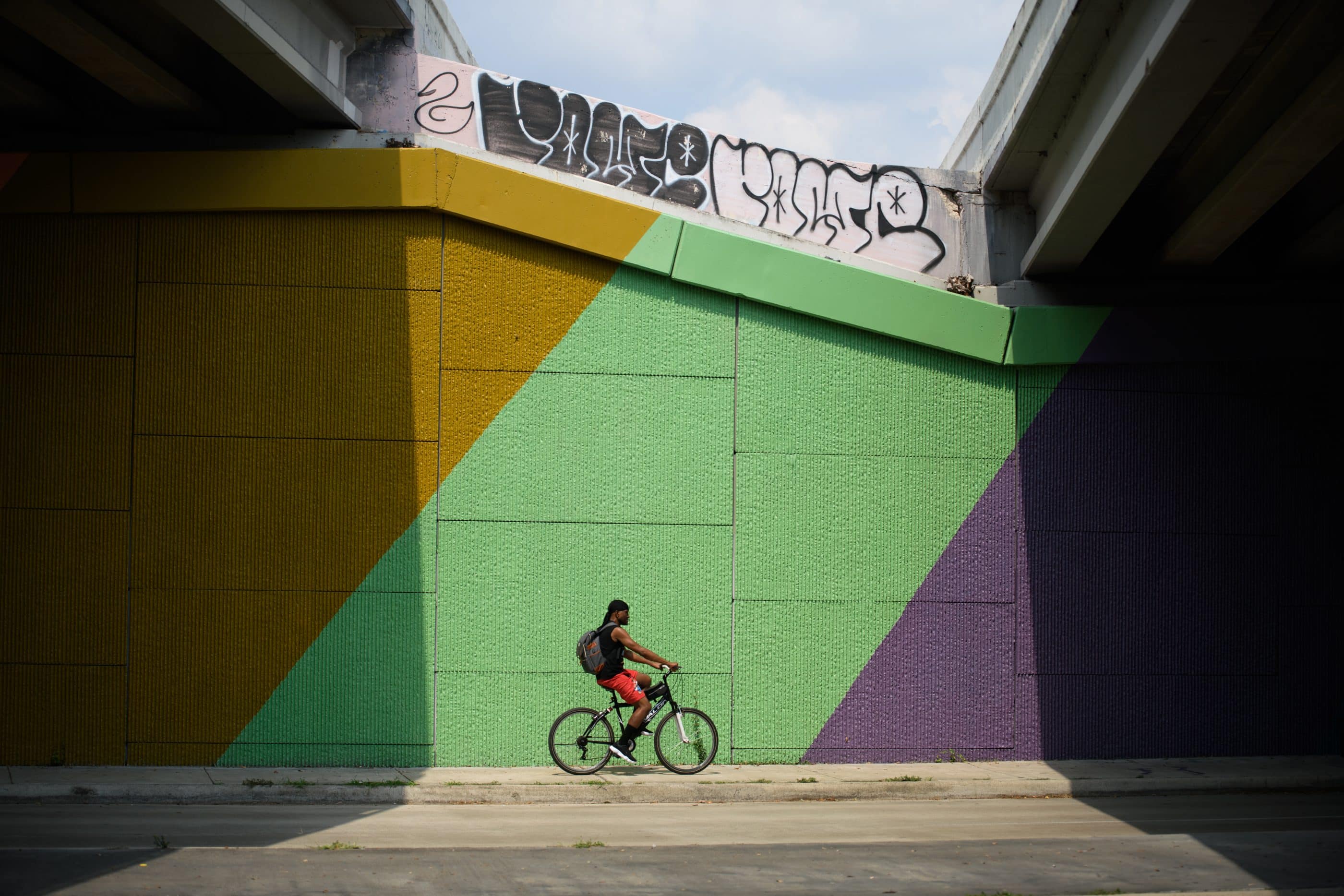 A bicyclist crosses in front of a mural in an underpass. The mural is made up of orange, green, and purple colorblocking.