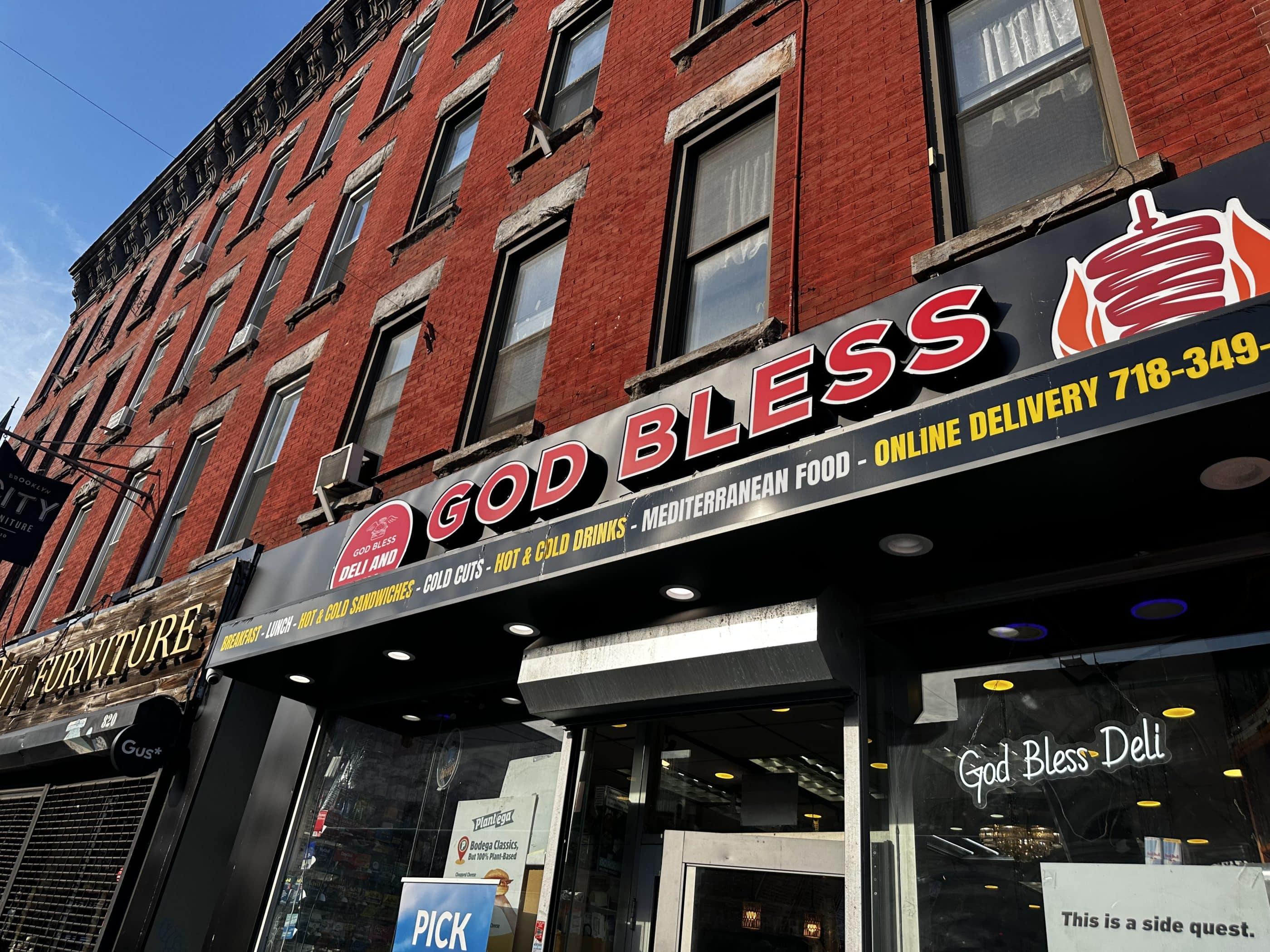 The front exterior of a bodega/deli in Brooklyn, New York. A large sign says GOD BLESS in 3D red-and-white capital letters. An awning advertises products sold at the deli, including cold cuts and Mediterranean food.