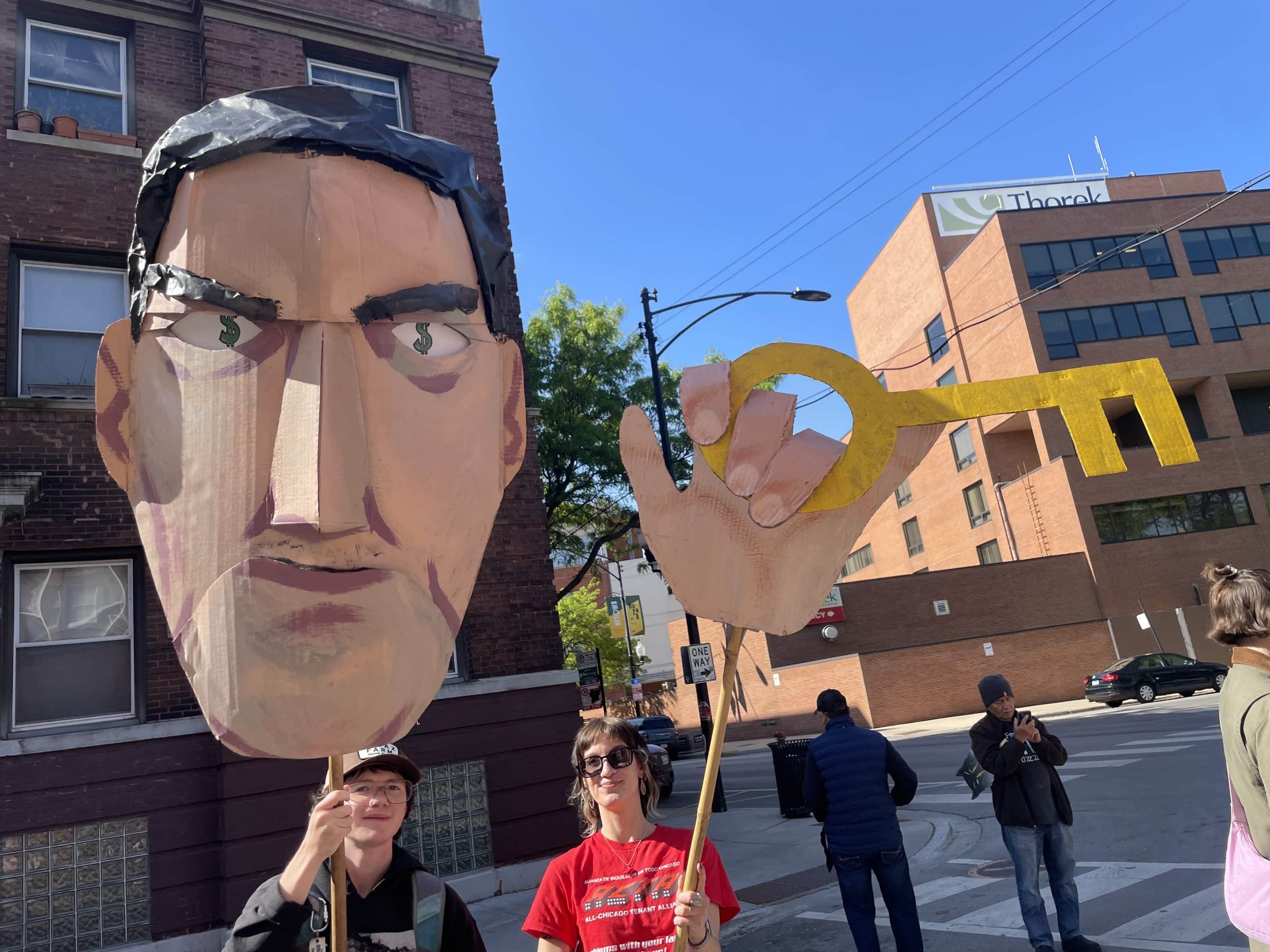 Two young white adults stand outside and hold up a large paper-mache head with dollar signs for eyes and a hand holding a golden key. One of them wears a red t-shirt that says All-Chicago Tenant Alliance.