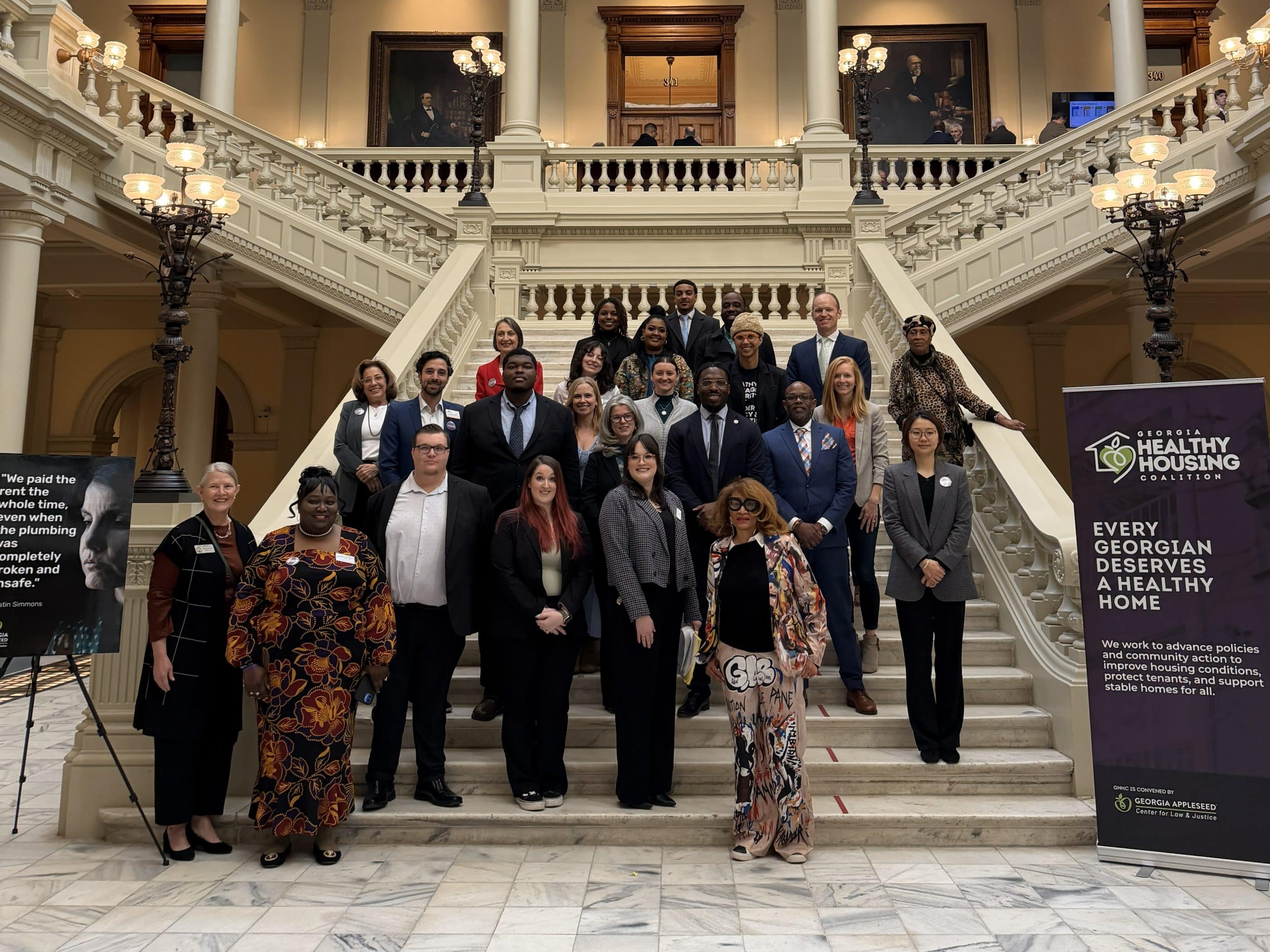 A large group of people of different races, ages, and genders, standing on a large marble staircase in a state capitol building. At the bottom of the staircase are two signs, one of which reads "Georgia Healthy Housing Coalition: Every Georgian deserves a healthy home."