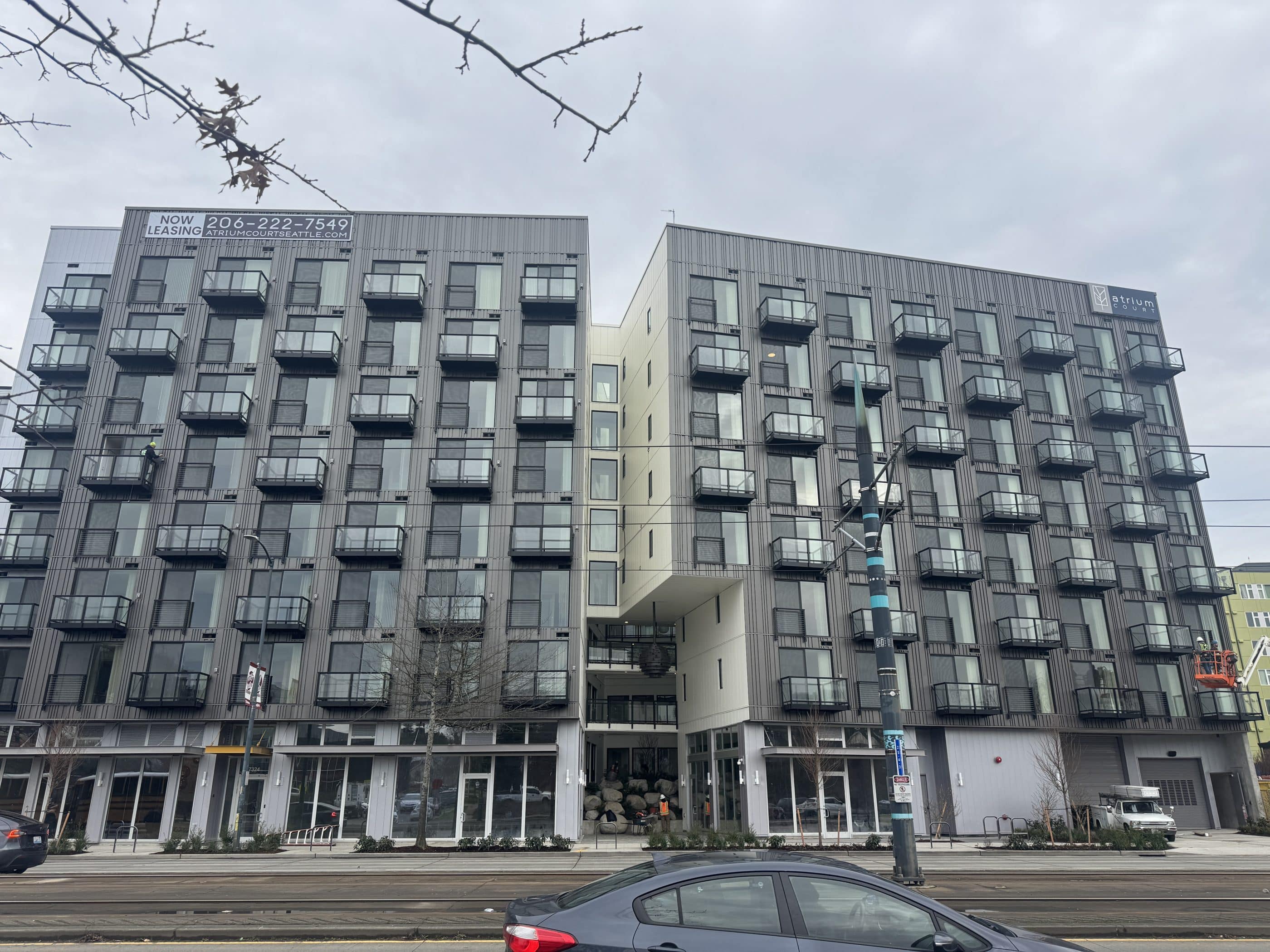 A large gray apartment building in an urban area, featuring many glass-and-iron front balconies and a sign that reads Atrium Court at the top of part of the building. A sign on another part of the building reads Now Leasing, with a telephone number and website for the building. Cars and tram tracks can be seen in the foreground.