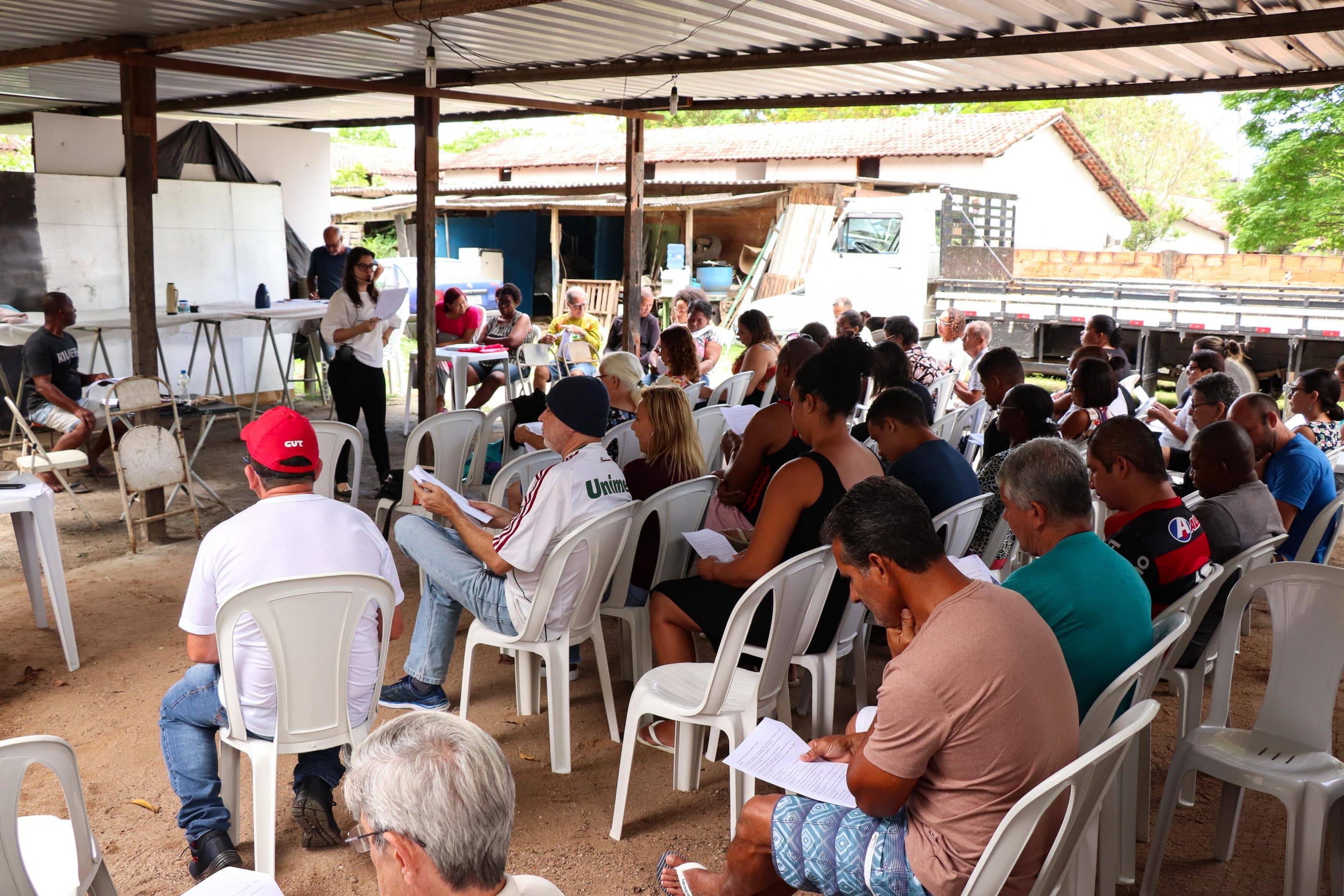 A large group of people sit on white plastic chairs under a partially covered outdoor structure. A woman stands at the front of the space, reading from a sheet of paper. She wears a microphone and appears to be reading something to the crowd.