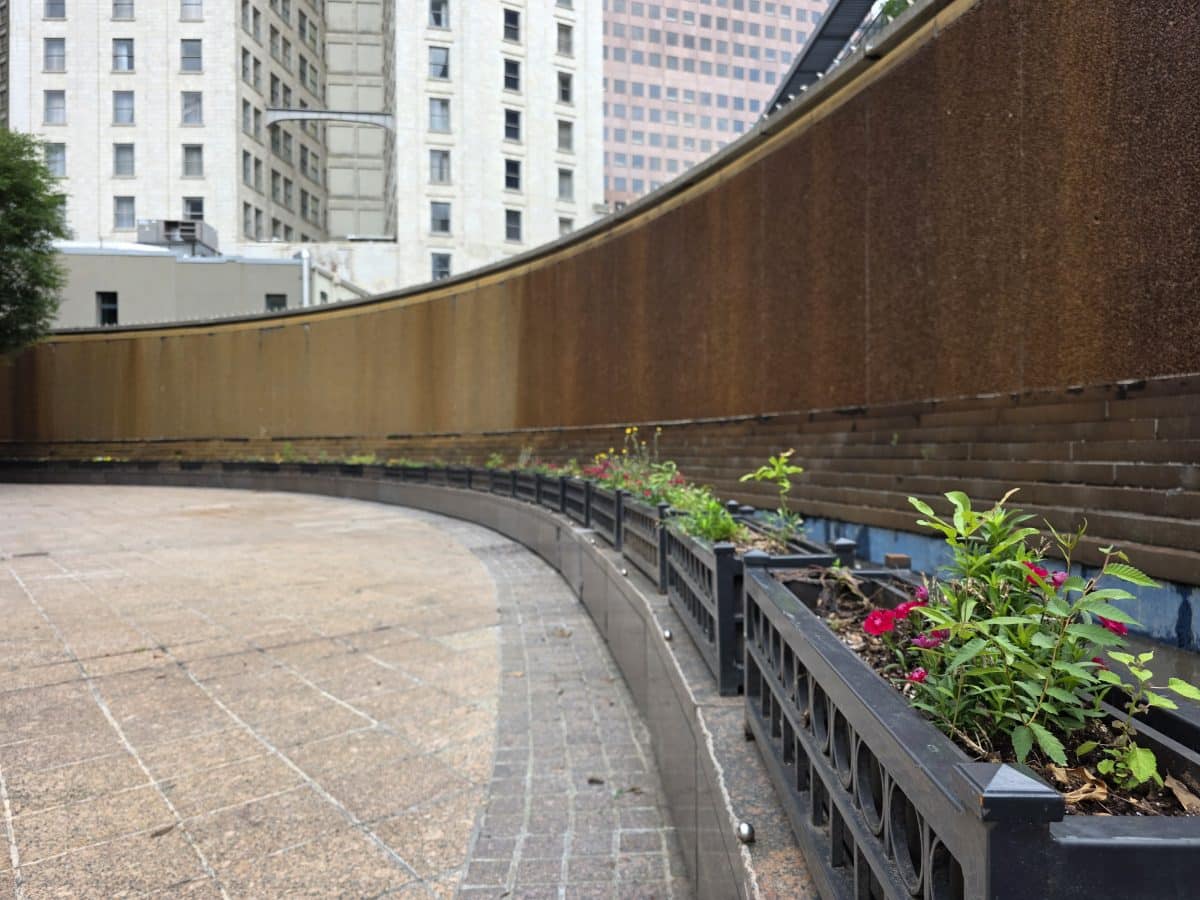 A curved outdoor water feature/wall in a public park. Planters with pink and yellow flowers and green grass follow the curve of the water feature.