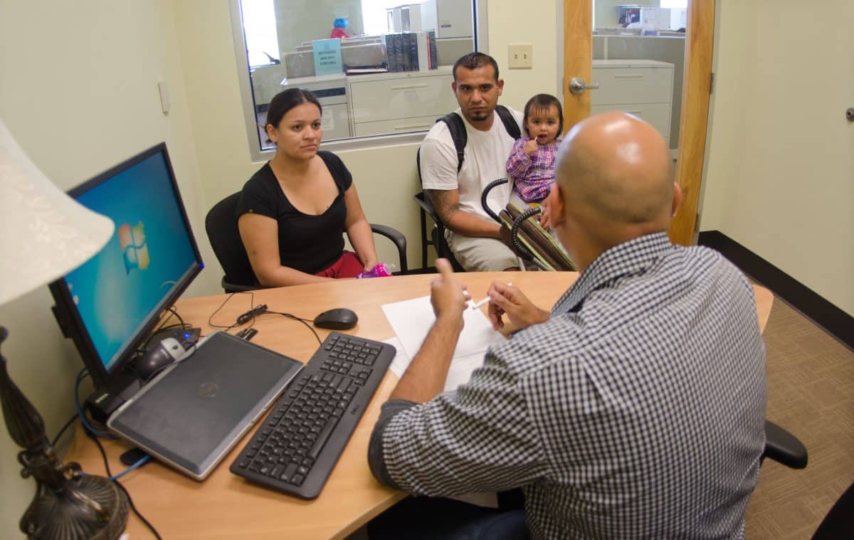 A casually dressed young man and woman sit with their toddler in a small office room. A man with his back to the camera sits at a desk in front of them, with paperwork on the table.