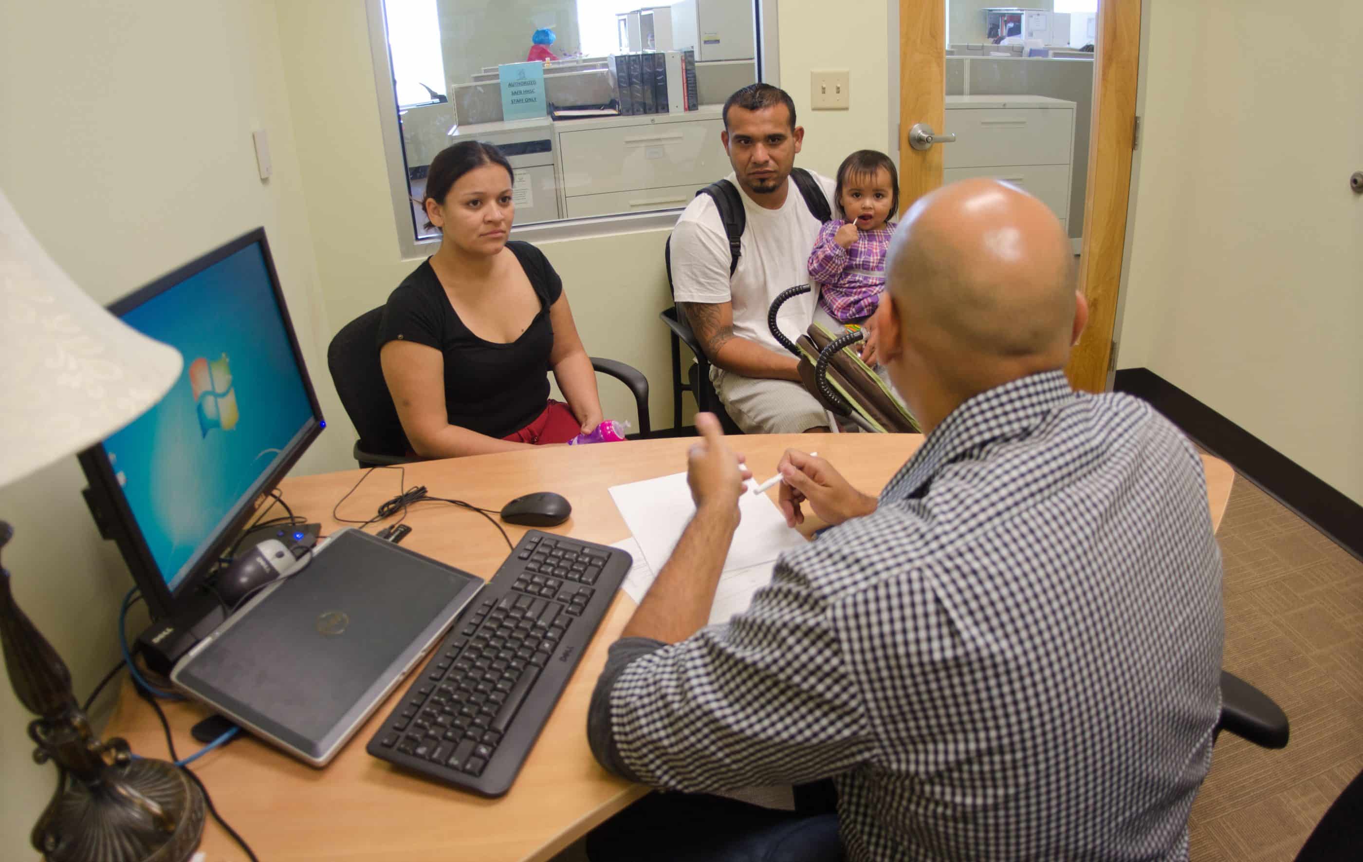 A casually dressed young man and woman sit with their toddler in a small office room. A man with his back to the camera sits at a desk in front of them, with paperwork on the table.