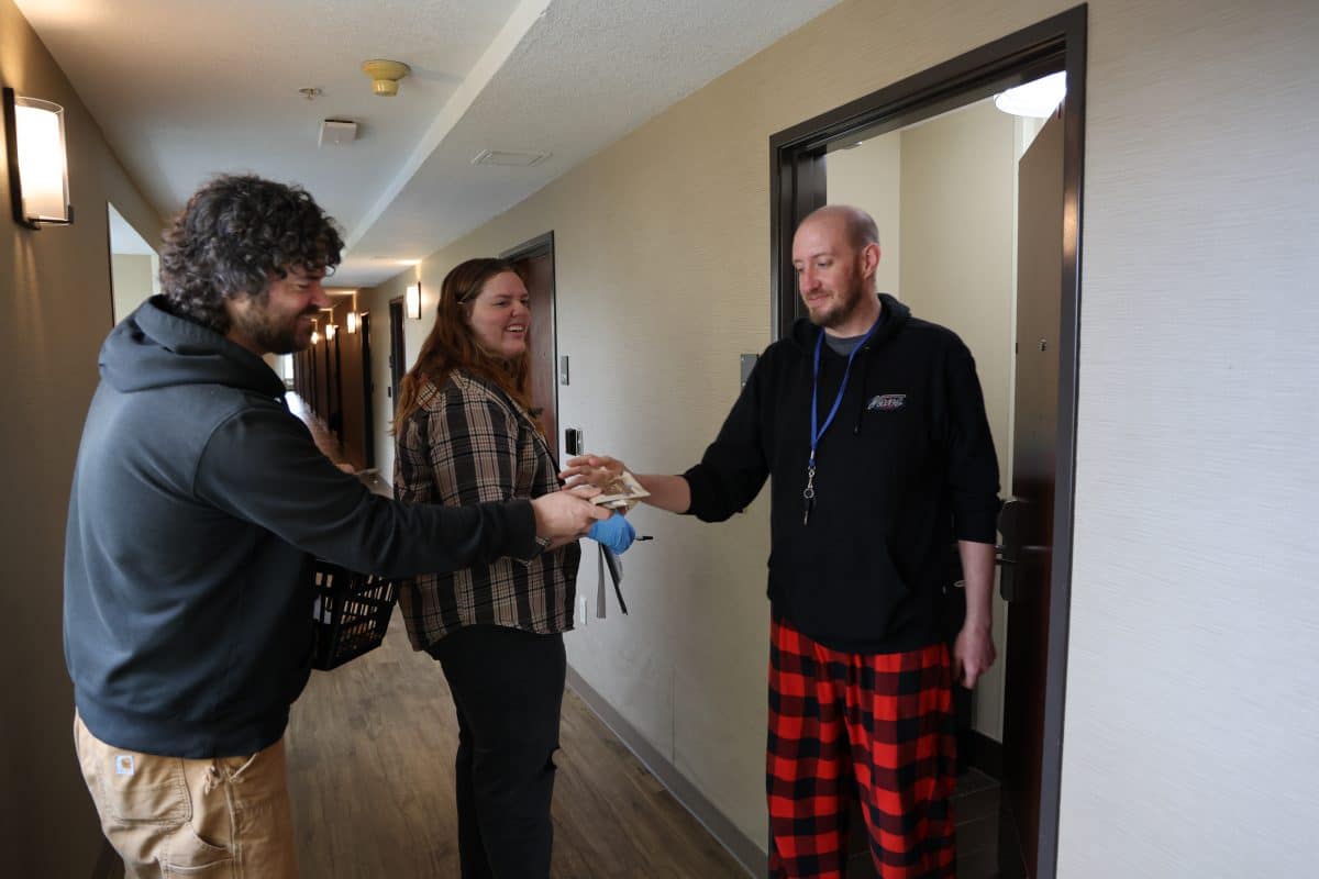 A white man with curly hair and a short beard, wearing a black sweatshirt and tan Carhartt pants, hands supplies to a white man with a close-shaved head and short beard, wearing a black Vans sweatshirt, and checkered red-and-black pajama pants. They are standing in the interior doorway of an apartment in what appears to be a residential building. A white woman with strawberry-blonde hair, wearing a checkered shirt and dark pants, stands behind them, holding a pen and papers in her hands.