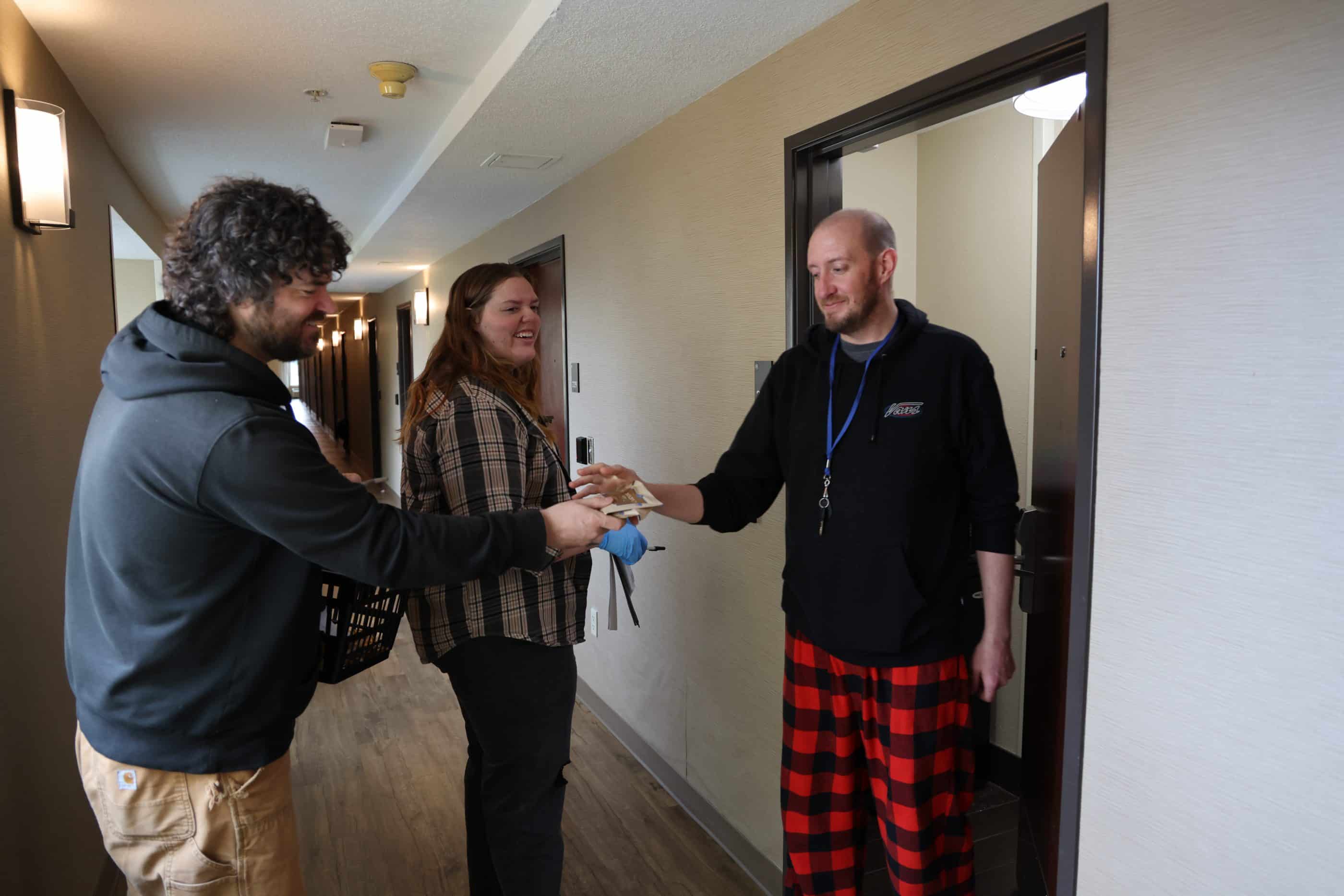 A white man with curly hair and a short beard, wearing a black sweatshirt and tan Carhartt pants, hands supplies to a white man with a close-shaved head and short beard, wearing a black Vans sweatshirt, and checkered red-and-black pajama pants. They are standing in the interior doorway of an apartment in what appears to be a residential building. A white woman with strawberry-blonde hair, wearing a checkered shirt and dark pants, stands behind them, holding a pen and papers in her hands.