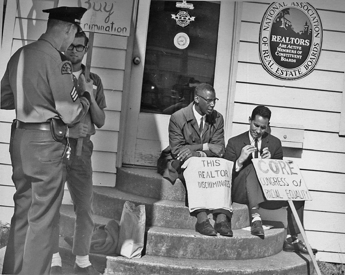 Two men sit on the front steps of a National Association of Real Estate Boards building, holding signs that read "This realtor discriminates" and "CORE: Congress of Racial Equality." A policeman standing off to the left talks with a man holding a protest sign.