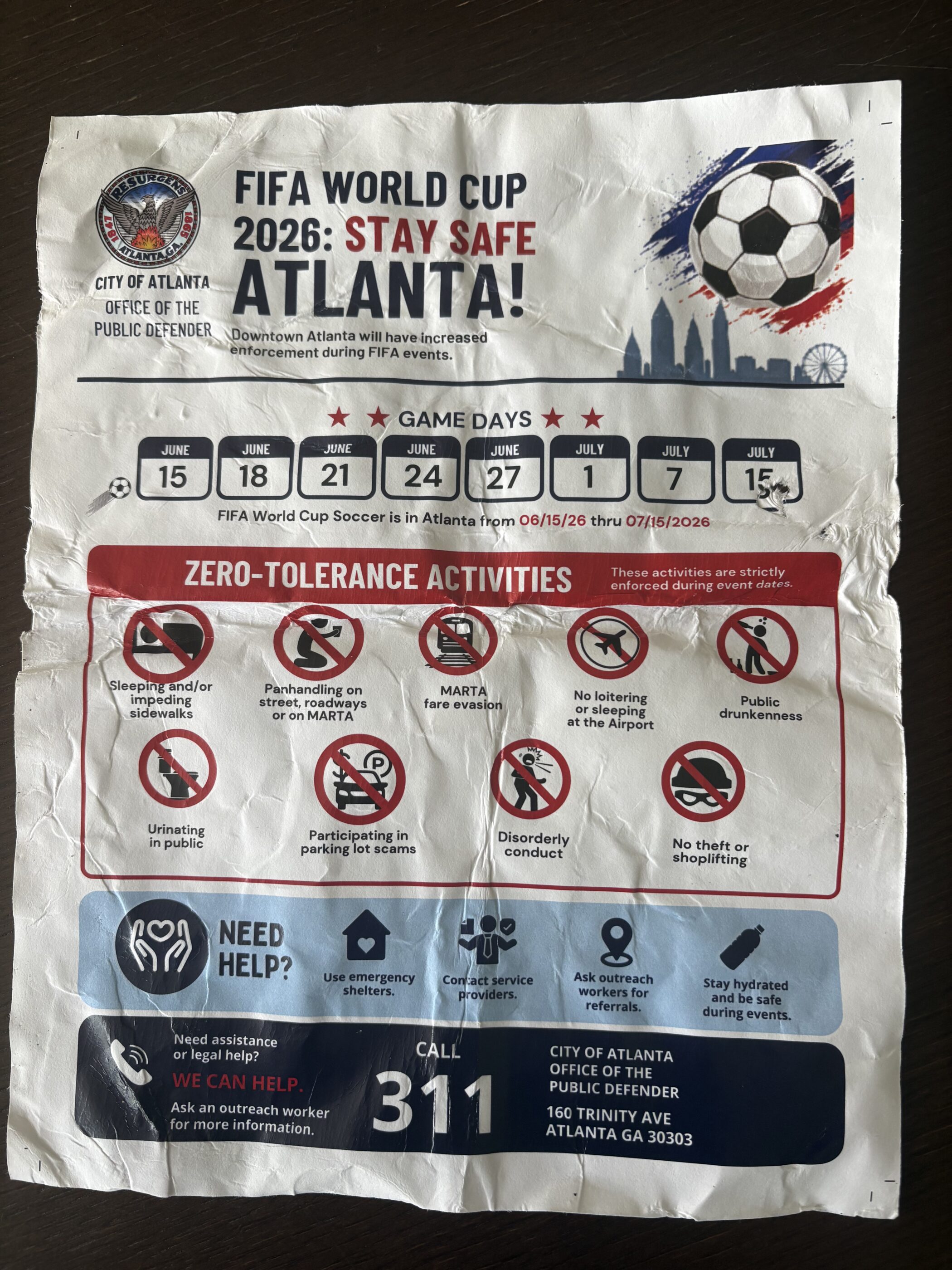 A wrinkled flyer reading "FIFA WORLD CUP 2026: STAY SAFE ATLANTA!" The flyer features information about "zero-tolerance activities," including panhandling, urinating in public, loitering, and sleeping on and/or impeding sidewalks. A City of Atlanta Office of the Public Defender logo adorns the top of the flyer.