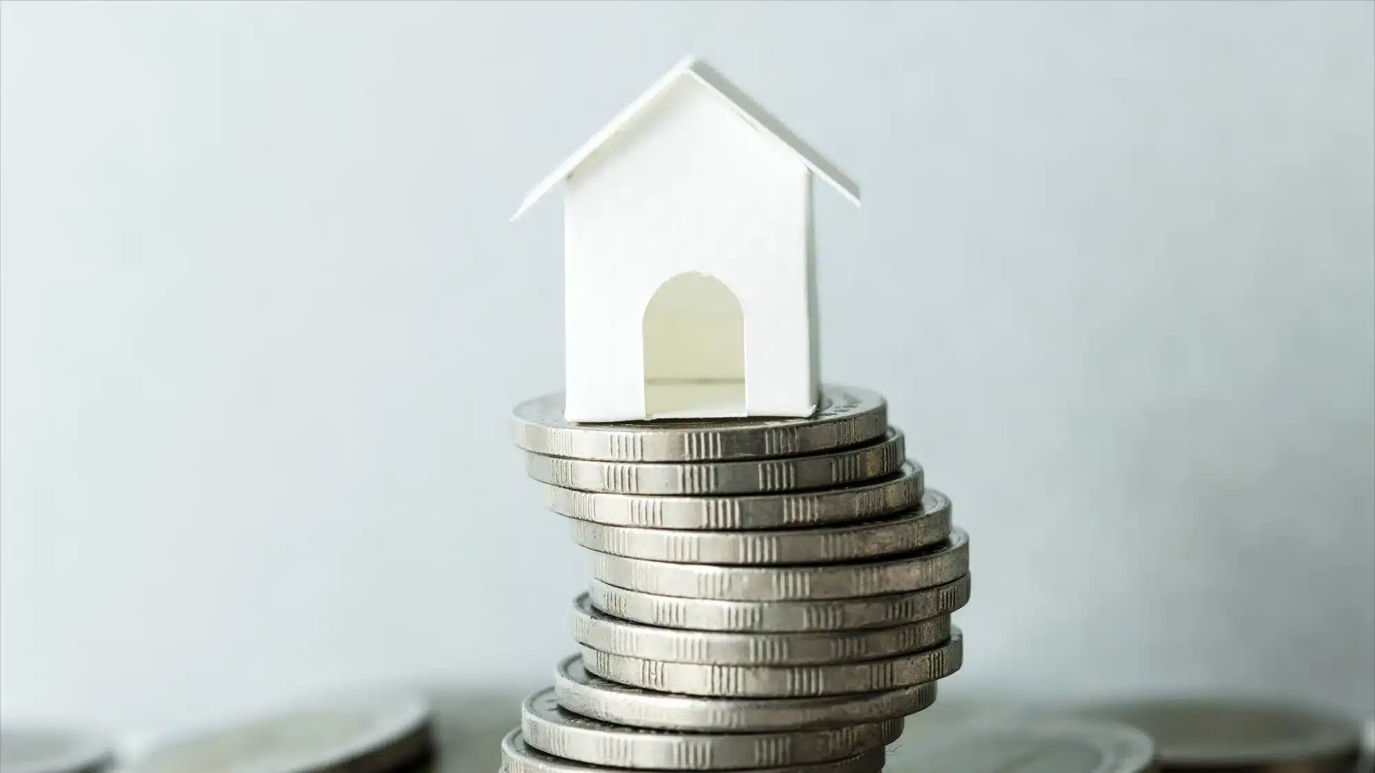 A small white house made out of paper sits atop a pile of silver coins.