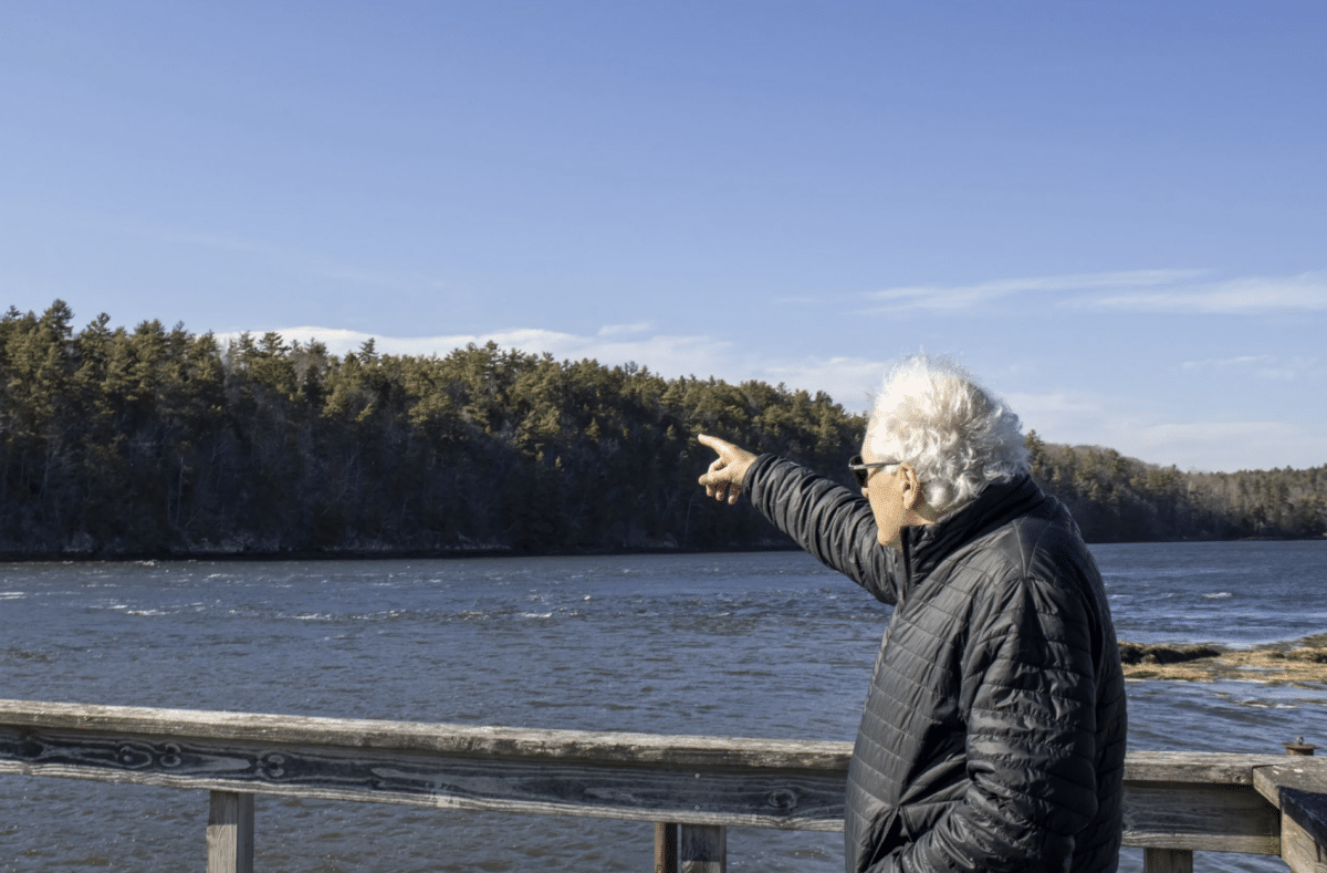 A white man with gray hair, wearing a black puffer jacket, stands on a dock overlooking a body of water bordered by tall trees. He points into the distance with his right arm.