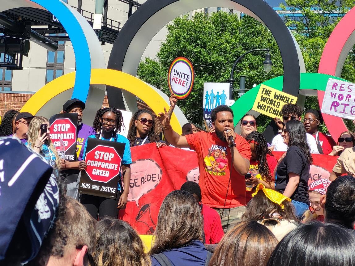 A diverse but majority Black crowd stands in front of an Olympic rings statue with various pro-labor signs such as "Stop Corporate Greed" and "UNITE Here." A man in an red-orange shirt holds a microphone.