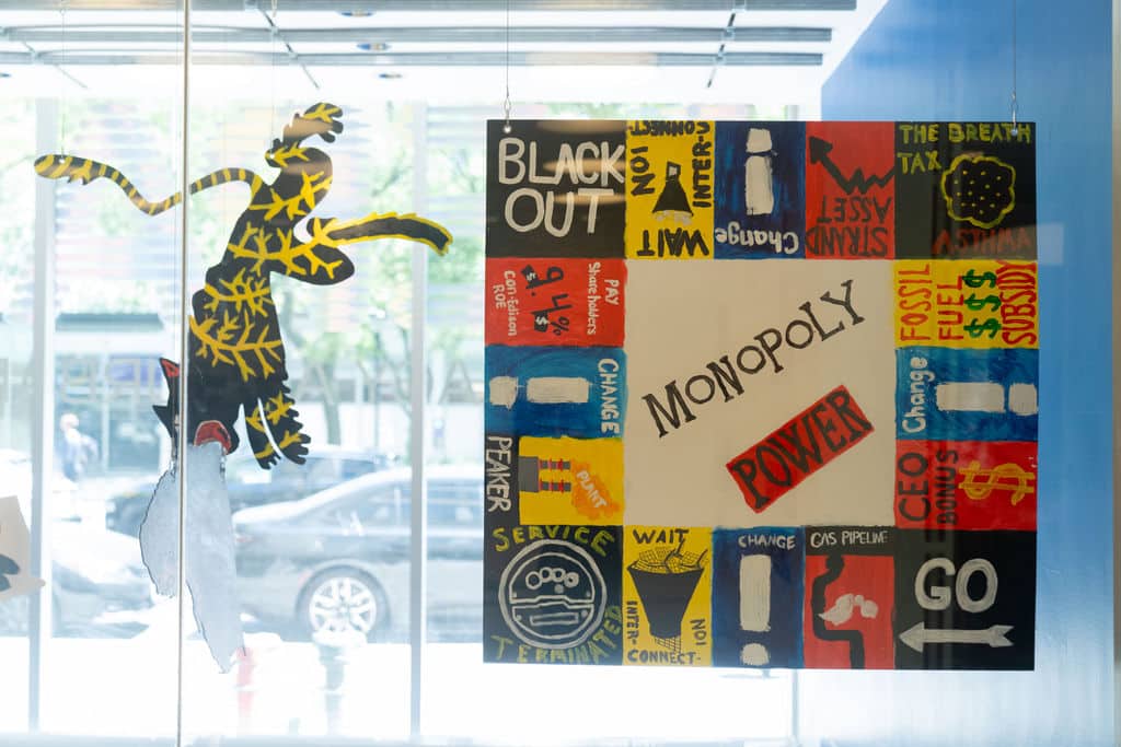 Two painted shapes are suspended in a window: a monopoly board with squares related to the power system, and a large cat painted with lightning bolts
