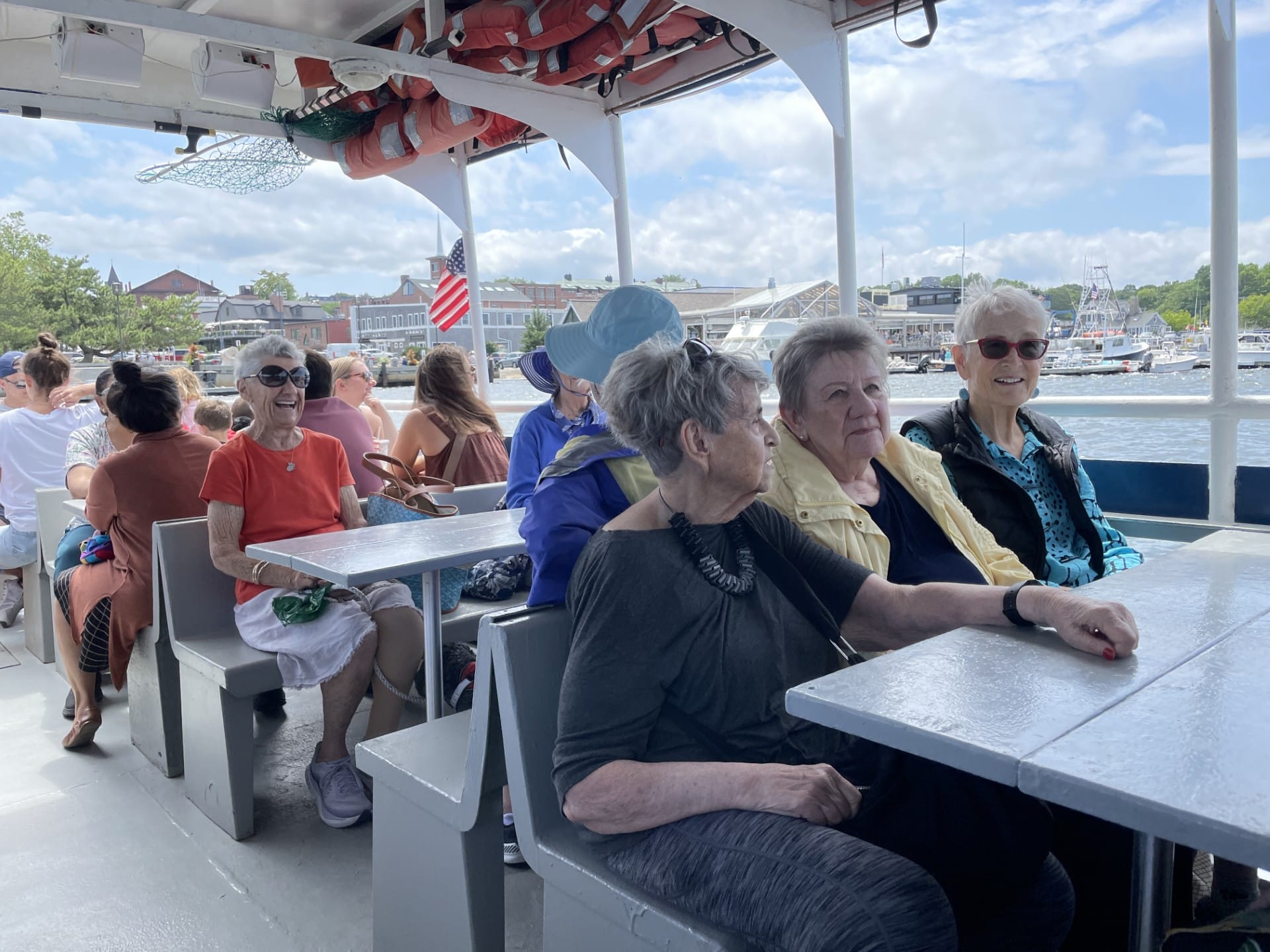 A group of older women sit at tables on a harbor cruise ship. Lifejackets and nets hang from the roof of the boat. Other boats and a variety of buildings are shown in the background.