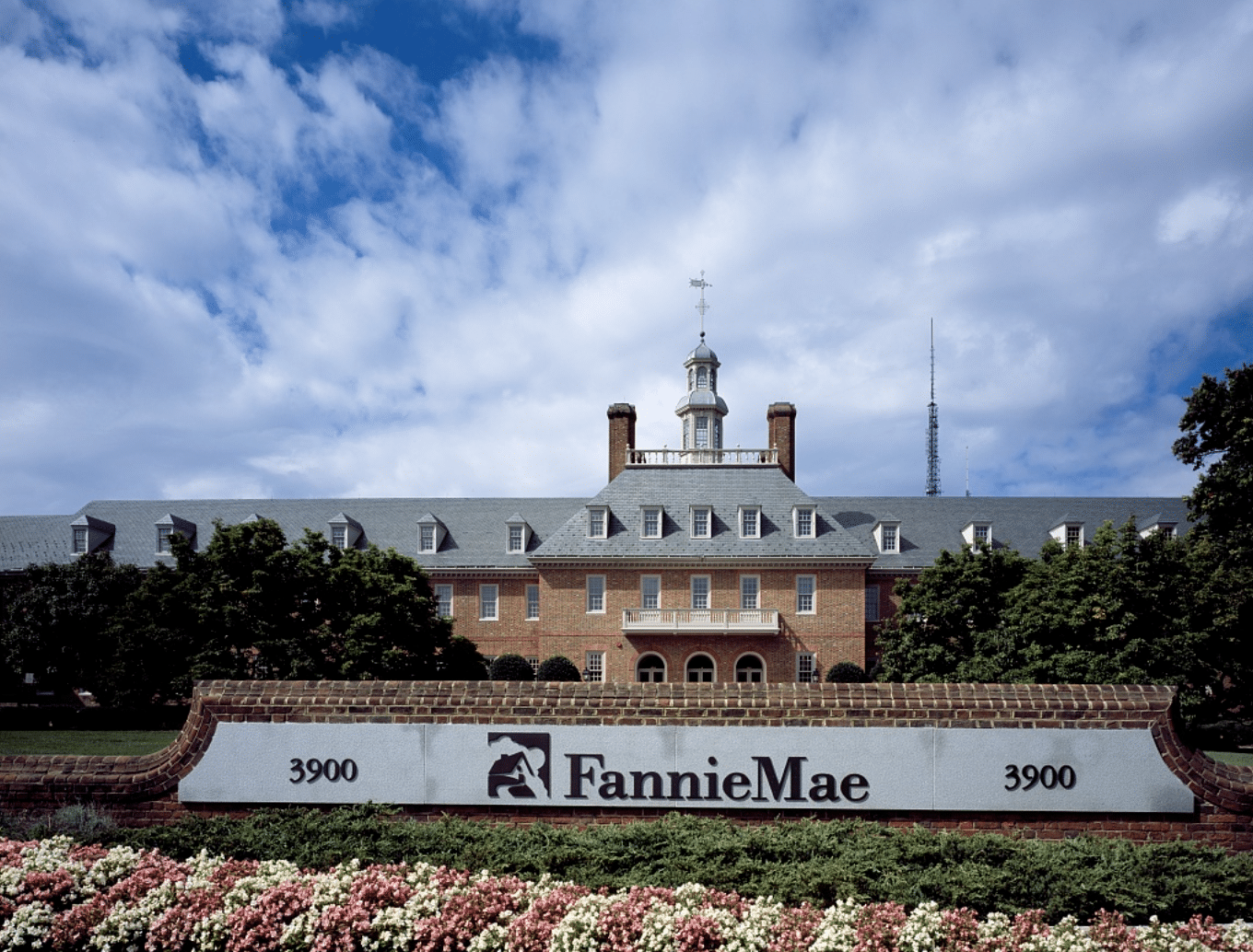 A large brick building with a spire and front balcony sits behind a large brick sign with FannieMae written on it. Pink and white flowers, along with green shrubbery, are planted in front of the sign.