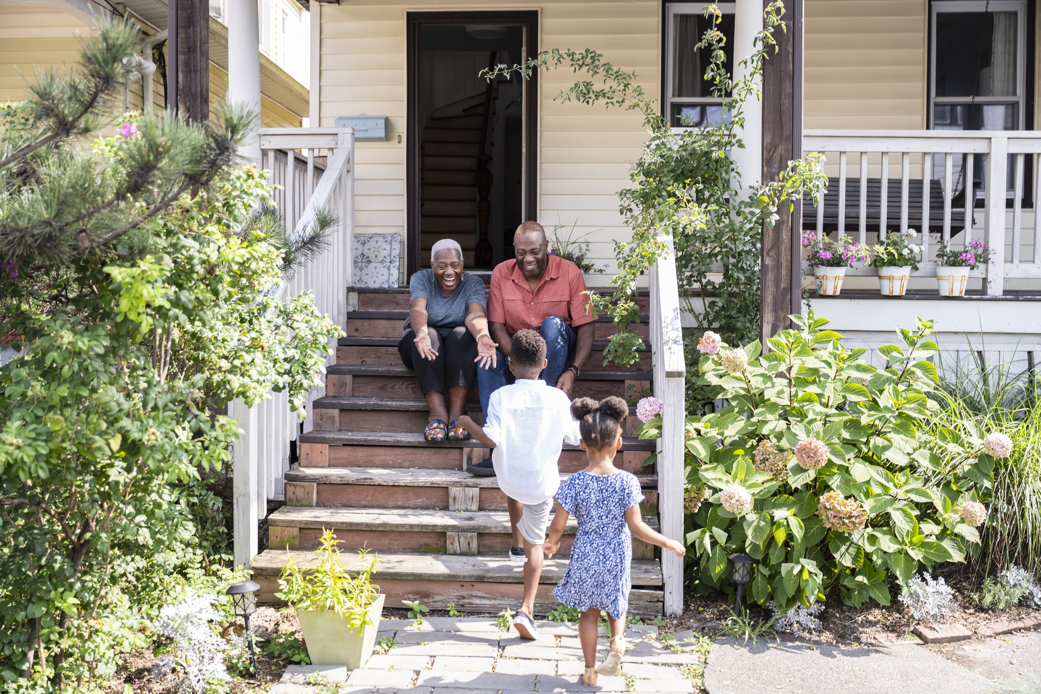 An older Black woman and an older Black man sit on the front steps of a yellow house. Two young Black children—a boy and a girl—walk up the stairs toward them. The woman holds out her arms to the children.
