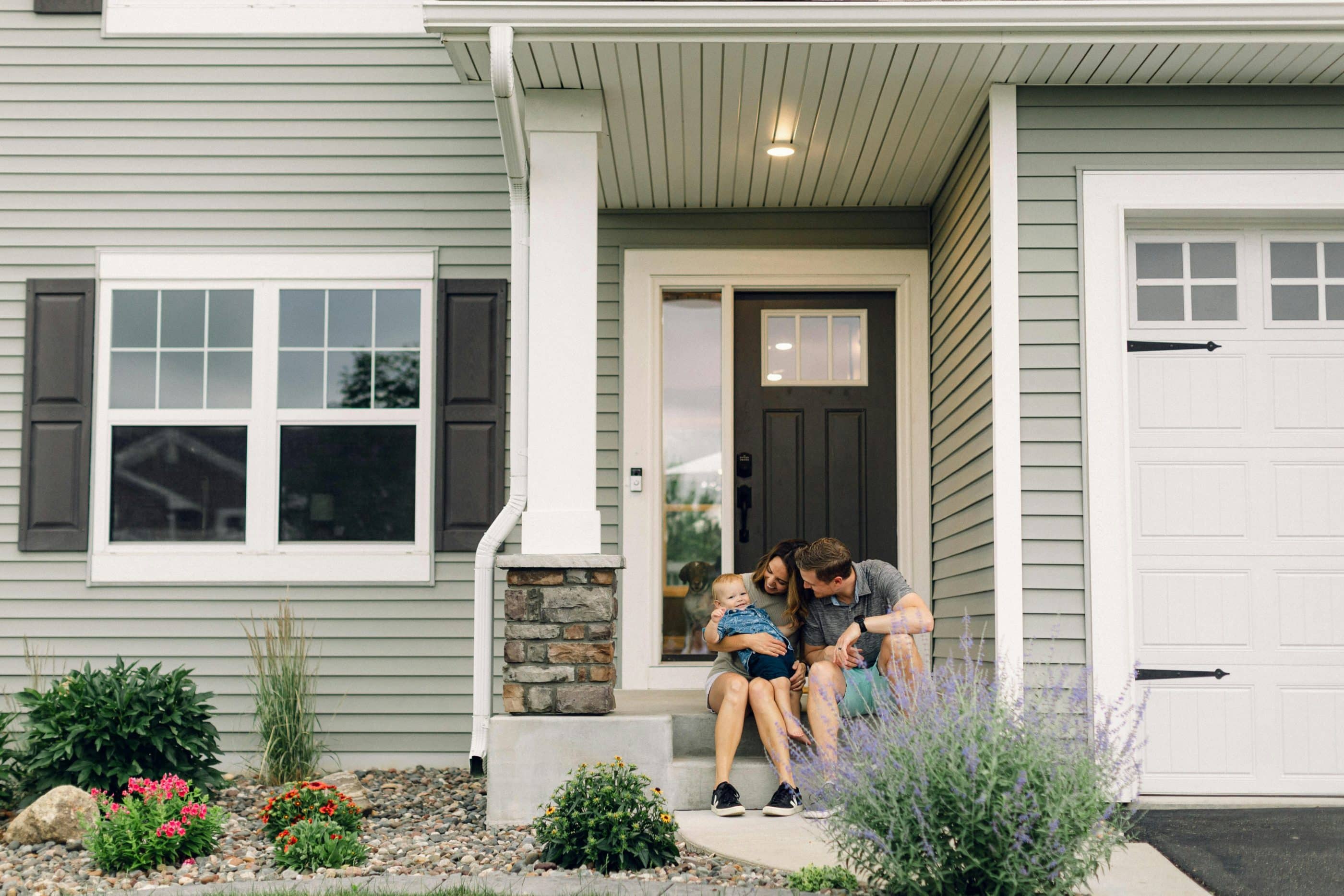 A young white couple—a man and a woman—wearing summery clothing sit on the front steps of their home with their infant son. Behind them is the front door of their house, where a dog can be seen peeking behind a door side window. The family's house is light green, with white windows and black shutters. A landscaped area of short plants and pebbles is in the foreground.